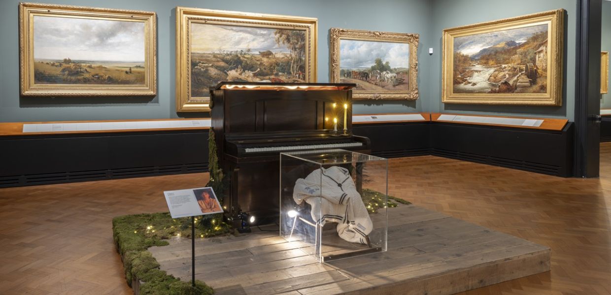 Installation shot of Taylor Swift Songbook Trail at the V&A. A cardigan in a glass box and piano in a room full of paintings. 